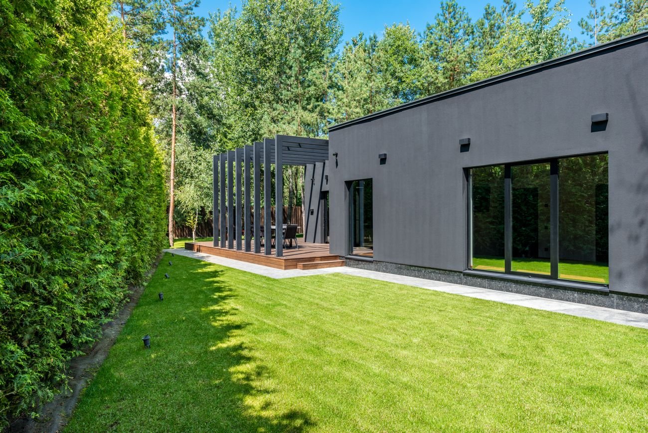Jardín residencial con césped verde, un seto y una terraza junto a la casa.